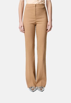 Pantaloni beige a vita alta e gamba dritta in tessuto texturizzato. Presentano una chiusura con zip frontale e due tasche laterali. Indossati con scarpe argentate.