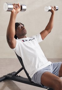 Persona levantando pesas plateadas y brillantes en un gimnasio de colores claros. Viste una camiseta blanca con un logo y pantalones cortos grises con un patrón, sentado en un banco negro.
