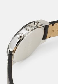 Montre en acier inoxydable argenté avec une lunette texturée noire et un bracelet en cuir. Comprend des poussoirs doubles et un fond de boîtier lisse.