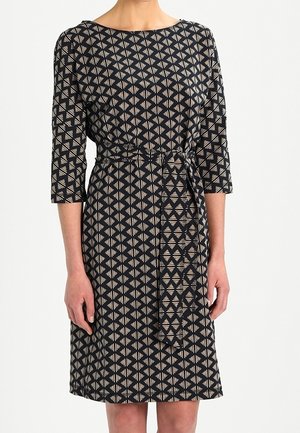 Kvinde iført en knælængde sort kjole med beige geometriske diamantmønstre og trekvartærmer, bundet med et matchende stofbælte.