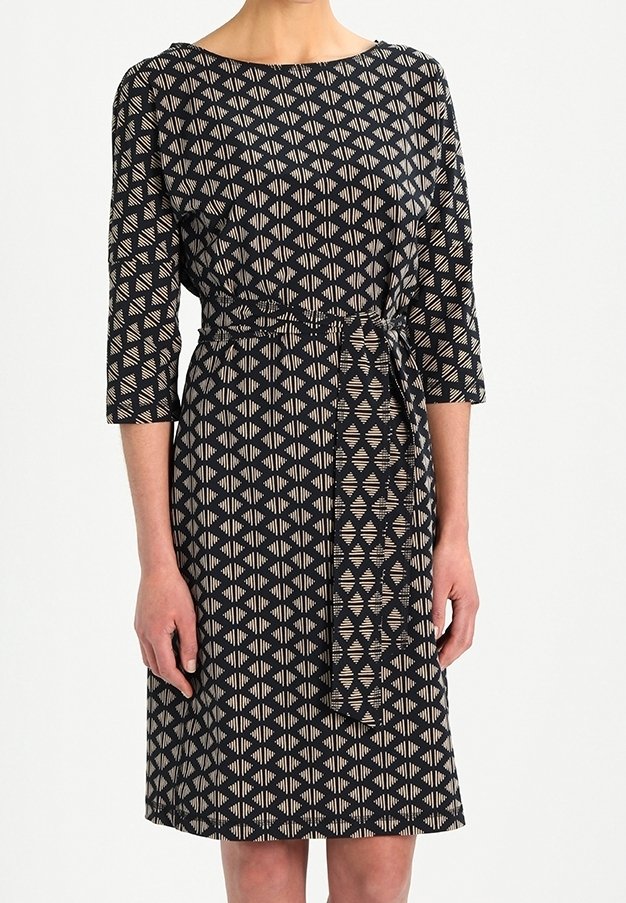 Femme portant une robe noire mi-longue avec des motifs géométriques beiges en forme de losange et des manches trois-quarts, ceinturée d'une ceinture assortie en tissu.