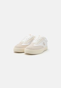White and beige sneakers with suede and fabric materials, rounded shape, thick rubber soles, and a small brand logo on the side.