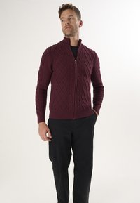 Burgunderfarbener, gestrickter Reißverschluss-Cardigan mit einem strukturierten Rautenmuster, geripptem Halsauschnitt, Bündchen und Saum, kombiniert mit schwarzen Hosen.