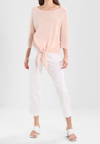 Top rosa chiaro a maniche lunghe con orlo annodato, abbinato a pantaloni bianchi a metà polpaccio. Il modello indossa tacchi bianchi con punta aperta. Tessuto dalla texture liscia.