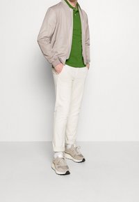 Veste bomber beige clair sur un polo vert, associée à un pantalon blanc et des baskets beiges. Matériau doux et coupe décontractée, avec des poignets retroussés.