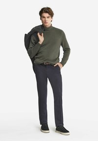Khaki golf z golfem, granatowe chinosy i czarne buty. Model trzyma szary płaszcz, stojąc na jednolitym białym tle.