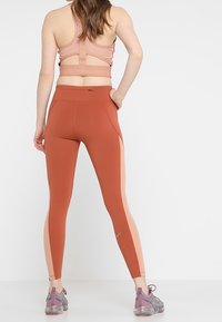 Top sportivo e leggings a vita alta in ruggine con pannelli laterali rosa chiaro. Realizzati in tessuto elasticizzato e traspirante. Presentano una schiena con taglio a forma di apertura.