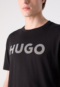 Melna kokvilnas T-kreklā ar izteiksmīgu "HUGO" logo tekstūrā pelēkā krāsā ar ziliem akcentiem. Īsas piedurknes un apaļa izgriezums.