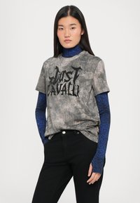 Hall gray t-särk musta "Just Cavalli" logoga, kantud sinise mustriga pikkade varrukatega pluusi peal, koos mustade pükstega. Tekstuuriga kangas ja mugav lõige.