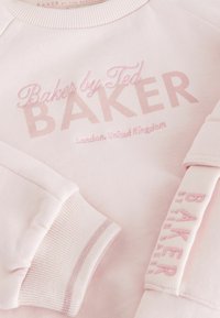 Gaiši rozā treniņtērps ar "Baker by Ted BAKE" logotipu tumšā rozā izšuvumā. Ar ribotiem piedurknēm un kaklu, ar mīkstu tekstūru.