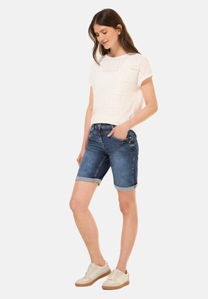 Haut blanc à œillets avec des manches courtes, associé à un short en denim foncé à revers. La modèle porte des baskets claires.