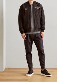 Giubbotto bomber nero con ricami gialli, maglione grigio, pantaloni scuri a motivi, e sneakers nere con suole bianche. Sfondo in legno.