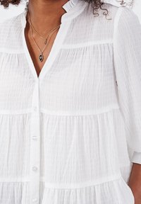 Blouse blanche texturée avec un col à volants, boutonnée à l'avant, et design à étages. Tissu semi-transparent avec manches longues légèrement froncées.