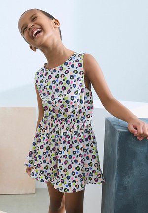 Enfant portant une robe sans manches colorée avec un motif léopard, riant et s'appuyant sur un bloc rectangulaire foncé.