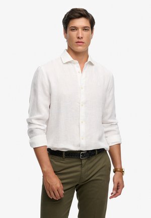 Joven con cabello oscuro que lleva puesta una camisa blanca de manga larga con botones, pantalones verde oliva, cinturón negro trenzado y un reloj de pulsera marrón.