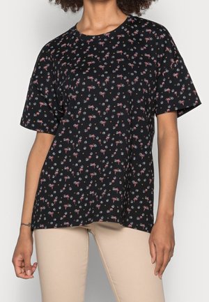 Kobieta ubrana w czarną koszulkę z krótkim rękawem z różowo-białym wzorem kwiatowym i beżowe spodnie, stoi z rękami swobodnie opartymi po bokach.