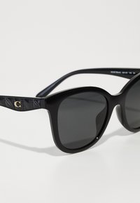 Lunettes de soleil noires avec une monture carrée audacieuse, dotées de verres sombres et de branches noires texturées, rehaussées d'un détail en logo doré.