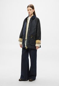 Tengerészkék vízálló dzseki arany díszítéssel, nagy zsebekkel és kontrasztos kockás szegéllyel. Világos pulóver fölött, sötét, bő szárú farmerrel viselve.