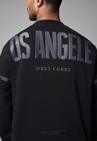 Čierna mikina s okrúhlym výstrihom a výrazným sivým textom "OS ANGELES" a "WEST COAST" na zadnej strane, s hladkou textúrou a minimalistickým dizajnom.