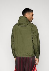 Ελαφρύ σακάκι σε ελιά πράσινο χρώμα με κουκούλα, ελαστικά περιλαίμια και τελείωμα. Ομαλή υφή, μίνιμαλ σχεδιασμός, ορατό από πίσω.