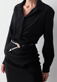 Blusa nera con colletto e maniche lunghe, caratterizzata da un dettaglio di tessuto drappeggiato. Abbinata a una cintura nera, accentata da una fibbia a forma di serpente.