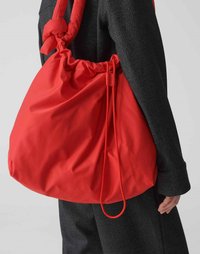 Borsa a tracolla rossa in nylon con cordoncino, caratterizzata da una superficie liscia e una base arrotondata, indossata su una giacca nera strutturata.