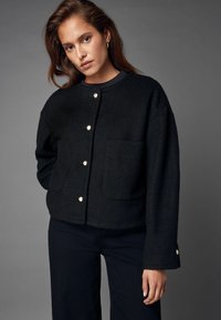 Schwarze, taillierte Wolljacke mit runder Halslinie, zwei vorderen Taschen und metallischem Knopfverschluss. Verfügt über lange Ärmel und eine weiche Textur.