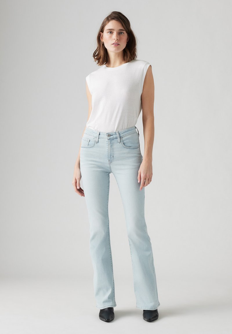 Jeans acampanados de color azul claro, hechos de denim, combinados con una blusa blanca sin mangas. El conjunto presenta una silueta ajustada y mínimos detalles de hardware.