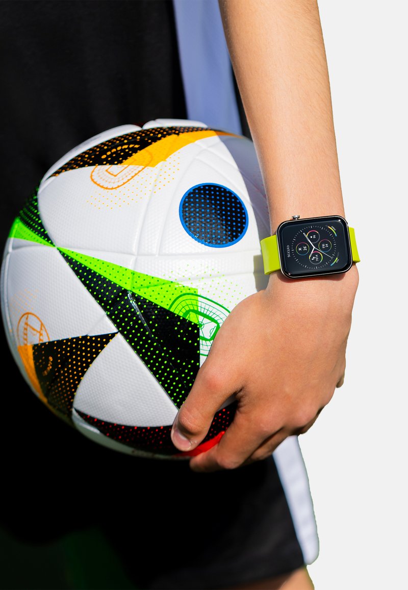 Vierkante smartwatch met een zwart horloge en een levendig groene band, vastgehouden terwijl er een veelkleurige voetbal met felle patronen en texturen wordt vastgegrepen.