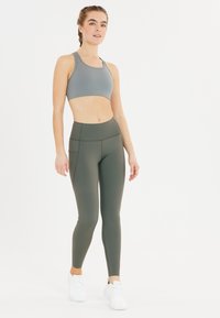 Lichtgrijze sportbeha gecombineerd met hoge olijfgroene leggings. De leggings hebben naden details en zijzakken. Witte sportschoenen.