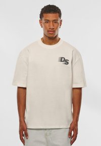 Cream-colored oversized t-shirt made of cotton, featuring short sleeves and a printed black logo on the left chest. Simple design with a round neck.