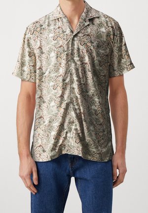 Homme portant une chemise boutonnée à manches courtes avec un motif géométrique abstrait beige, vert et marron et un jean bleu sur un fond uni.