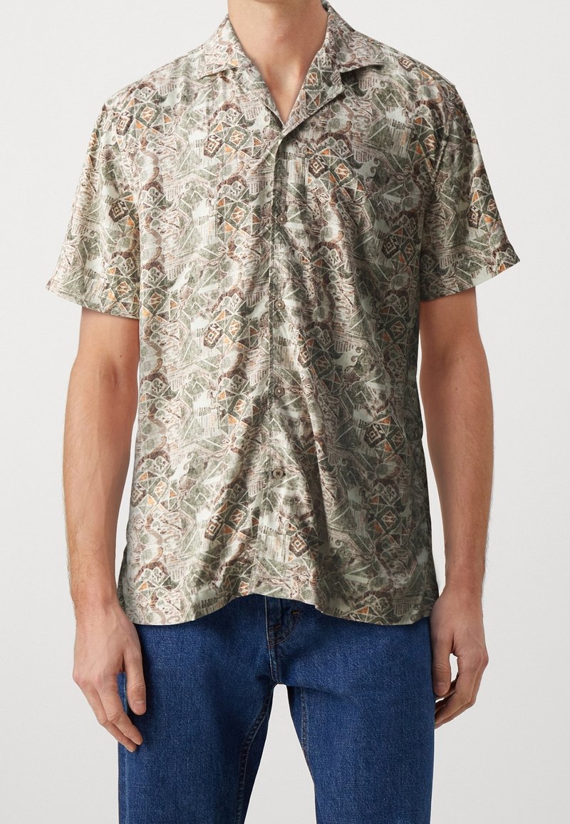 Homme portant une chemise boutonnée à manches courtes avec un motif géométrique abstrait beige, vert et marron et un jean bleu sur un fond uni.
