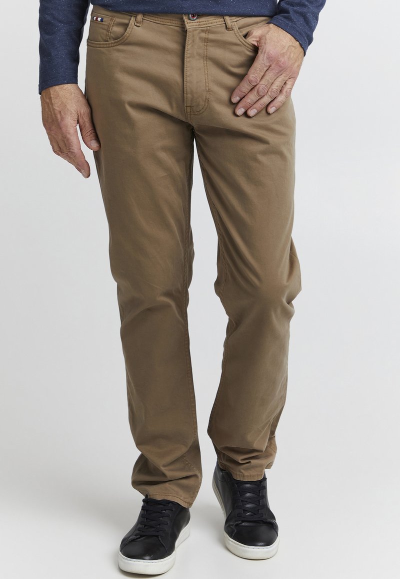 Braune Baumwollhose mit geradem Schnitt, ausgestattet mit einem Reißverschluss, Gürtelschlaufen und dezenten Nahtdetails. Kombiniert mit schwarzen Sneakers.