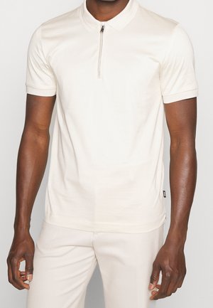 Man die een crème-kleurige korte mouwen polo draagt met een rits aan de voorkant en bijpassende lichtgekleurde broek tegen een effen achtergrond.