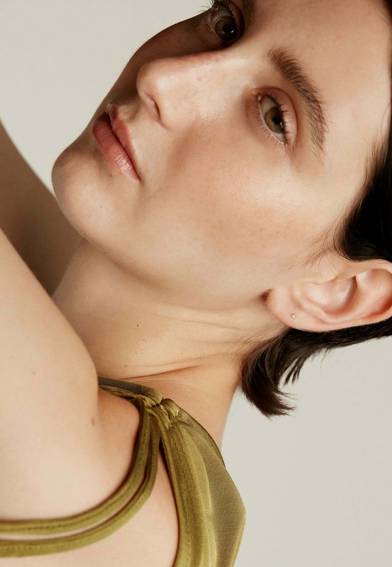 Top de color verde oliva con una tela suave, que presenta un delicado detalle de nudo en el hombro. Piel clara con sutiles pecas; arete de perno sencillo.