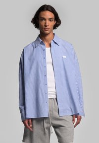 Camicia a righe blu e bianche con colletto classico, dettaglio della tasca sul petto e vestibilità rilassata, abbinata a pantaloni corti grigi.
