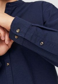 Man som fäster knapp på manschetten på en långärmad mörkblå skjorta som visar detaljerad tyggtextur och knappdesign.