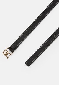Črn usnjen pas z belim šivom, z zlatom obarvanom kovinskim zaponko s podrobnostmi logotipa ter s tanko, ravno zasnovo.