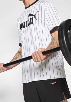 Personne portant une tenue de sport Puma blanche et noire soulève une barre avec des poids dans une salle de sport, montrant des avant-bras tatoués.
