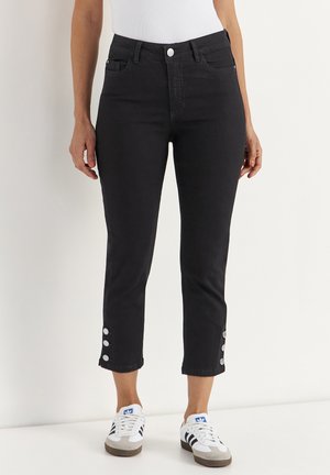 Kvinde iført sorte cropped skinny jeans med knapdetaljer nær anklerne, hvid top og hvide Adidas-sneakers med sorte striber.