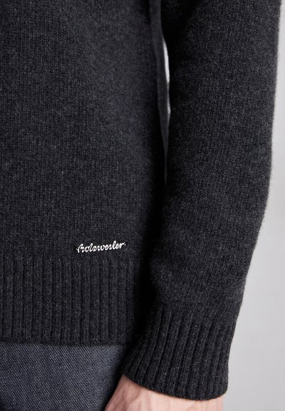 Grå strikket genser med ribbestrikket kant og mansjetter, med en liten hvit brodert logo nederst til venstre. Myk tekstur, tettsittende design.