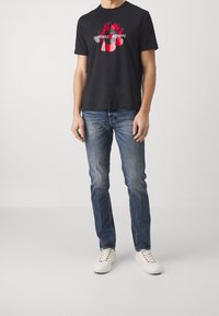 Svart bomullströja med en grafisk design i rött och svart med texten "Emporio Armani". Kombinerad med blå denimjeans och vita sneakers.