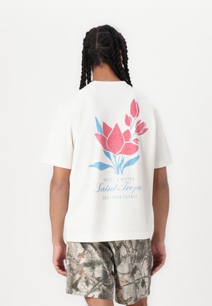 Person with braided hair wearing a white T-shirt featuring a pink and blue floral design and text, paired with camouflage-patterned shorts.