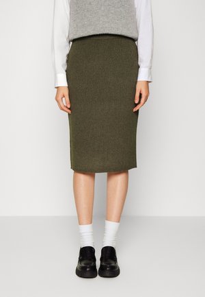 Person iført et olivengrønt ribbet blyantskørt, hvid langærmet skjorte, grå ærmeløs sweater, hvide sokker og sorte loafers, stående.