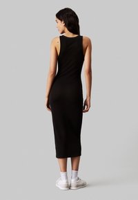 Abito midi nero a costine con design senza maniche, silhouette aderente e scollatura rotonda. Indossato con sneakers bianche e calzini alla caviglia.