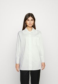 Camisa branca de botão com um grande colar texturizado, mangas longas e um bolso no peito. Feita de um tecido leve e suave.