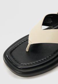 Beige synthetic flip-flop with a triangular toe post, featuring a textured black sole and curved design. Accented with stitching details.