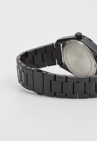 Melnā metāla rokas pulkstenis ar ķēdes aproci, apaļu korpusu un teksturētu ciparnīcu. Iekļauj kronīti, nerūsējošā tērauda aizmuguri un logo detalizāciju.