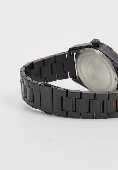 Montre en métal noir avec bracelet à maillons, boîtier rond et cadran texturé. Comprend une couronne, un dos en acier inoxydable et un détail de logo.